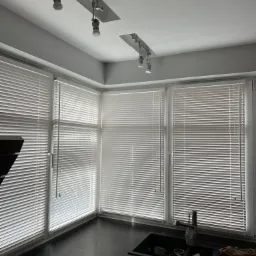 Wnętrze kuchni z trzema oknami zabezpieczonymi białymi żaluzjami poziomymi, oświetlonymi przez lampy sufitowe na szynoprzewodzie.