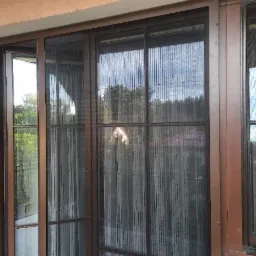 Zewnętrzna moskitiera plisowana w brązowej ramie, zamontowana na balkonie, widok na zieleń i domy w tle.