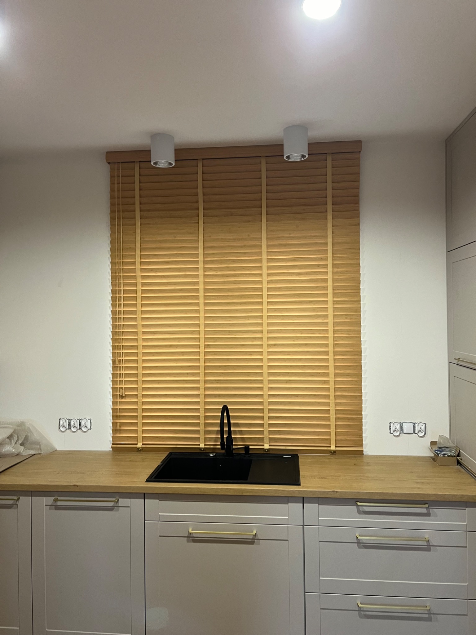 Jasne drewniane żaluzje poziome zamontowane w kuchni nad czarnym zlewem, widoczne białe lampy sufitowe i szare szafki kuchenne z złotymi uchwytami.