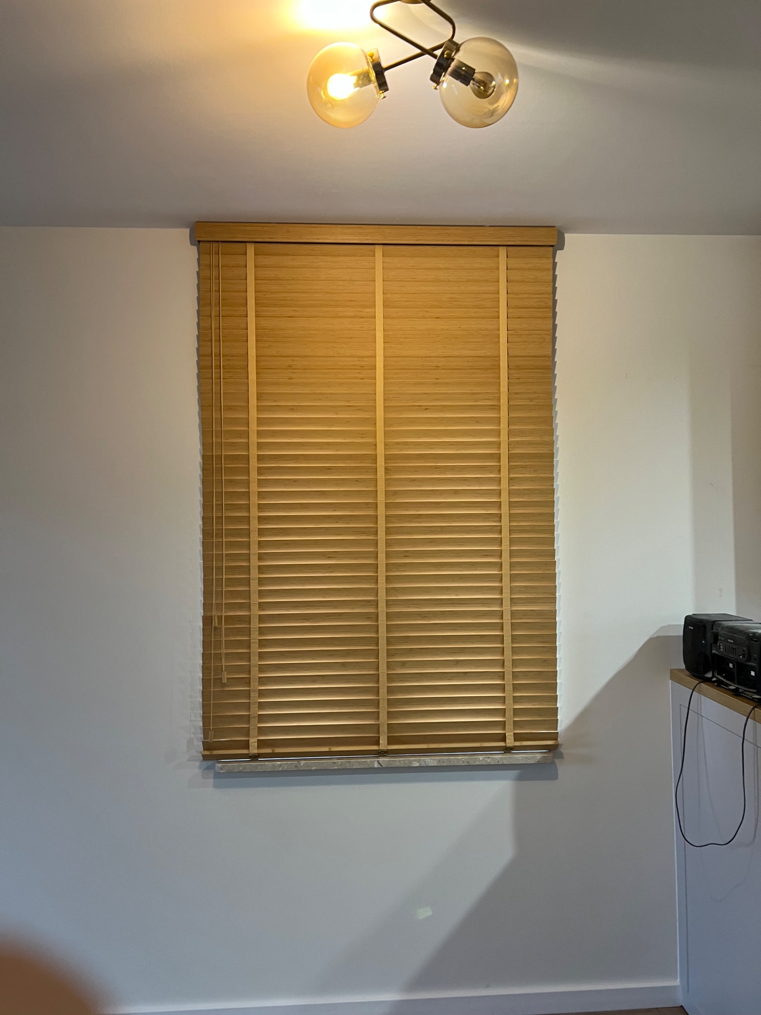 Zamknięte drewniane żaluzje w oknie, oświetlone ciepłym światłem wiszącej lampy, widoczne fragmenty sprzętu audio po prawej stronie.