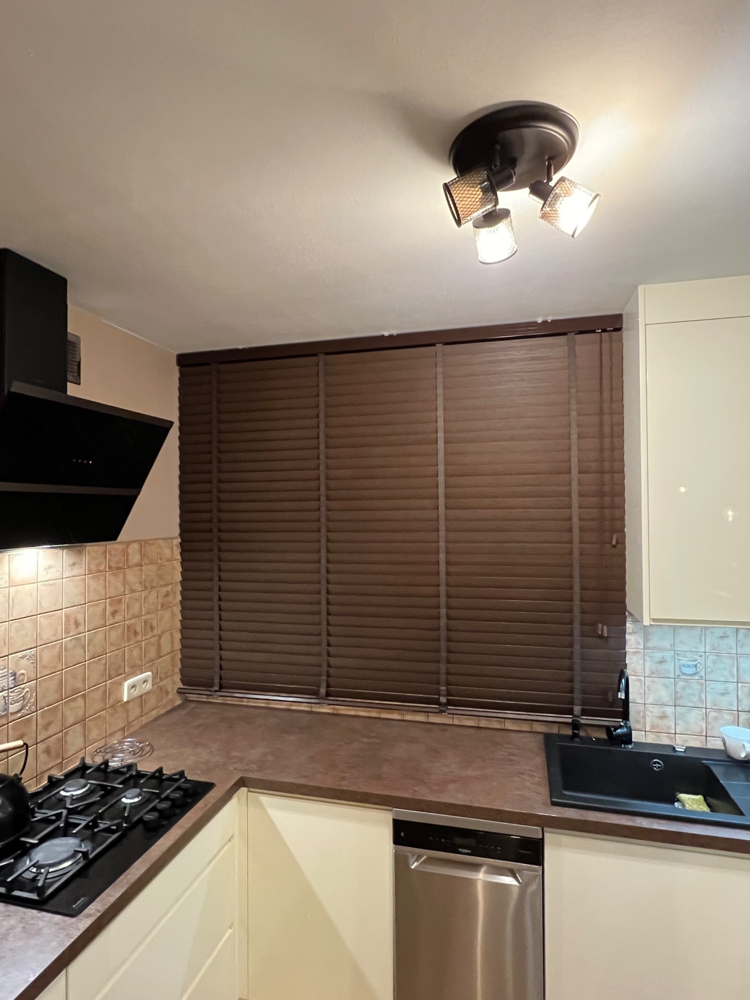 Kuchnia z brązowymi żaluzjami drewnianymi w oknie, widoczny fragment blatu, zlewu, płyty grzewczej i okapu.