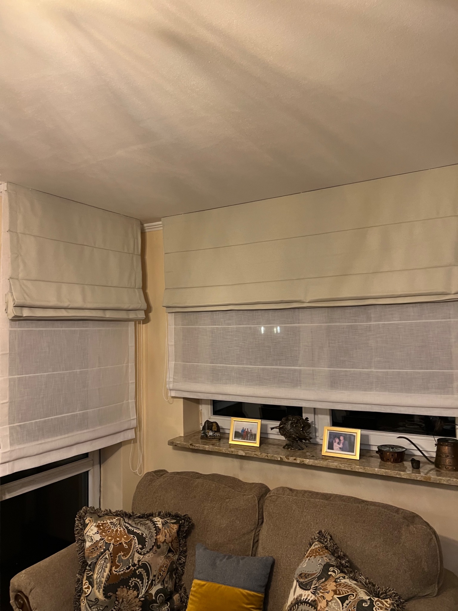 Dwa okna z zamontowanymi kremowymi roletami rzymskimi, częściowo opuszczonymi, z widocznymi sznurkami do regulacji, nad brązową sofą z dekoracyjnymi poduszkami, na parapecie zdjęcia w ramkach...