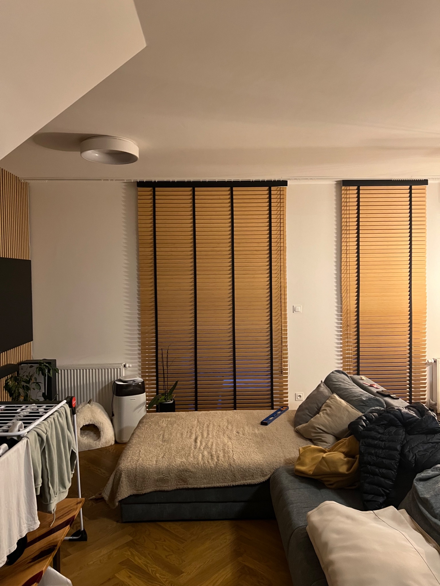 Jasne żaluzje drewniane z czarnymi elementami w nowoczesnym wnętrzu, widoczne z perspektywy pokoju dziennego z elementami codziennego użytku, takimi jak suszarka na pranie i rozłożona sofa.