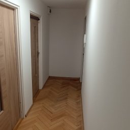 Usługi remontowo-budowlane Team Daniel's - Korytarz po remoncie: parkiet ułożony w jodełkę, świeżo pomalowane białe ściany, widoczne drzwi i okrągła lampa sufitowa. Jasne, nowoczesne wykończenie.