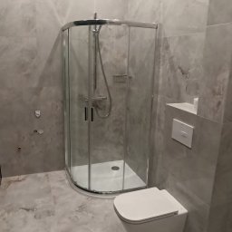 Usługi remontowo-budowlane Team Daniel's - Nowoczesna łazienka w szarościach: kabina prysznicowa z chromowanymi detalami, podwieszana toaleta, płytki imitujące marmur na ścianach i podłodze, minimalistyczny design.