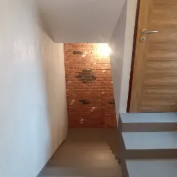 Schody wewnątrz budynku z szarą powierzchnią, ściany białe, fragment ściany wykończony czerwoną cegłą. Widoczne drzwi z drewnianą okleiną i srebrną klamką.