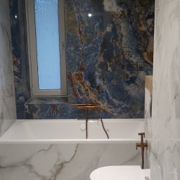 Usługi remontowo-budowlane Team Daniel's - Elegancka łazienka z marmurowymi płytkami, białą wanną i toaletą. Ściana za wanną wykończona płytkami imitującymi niebieski marmur. Armatura w kolorze miedzi.