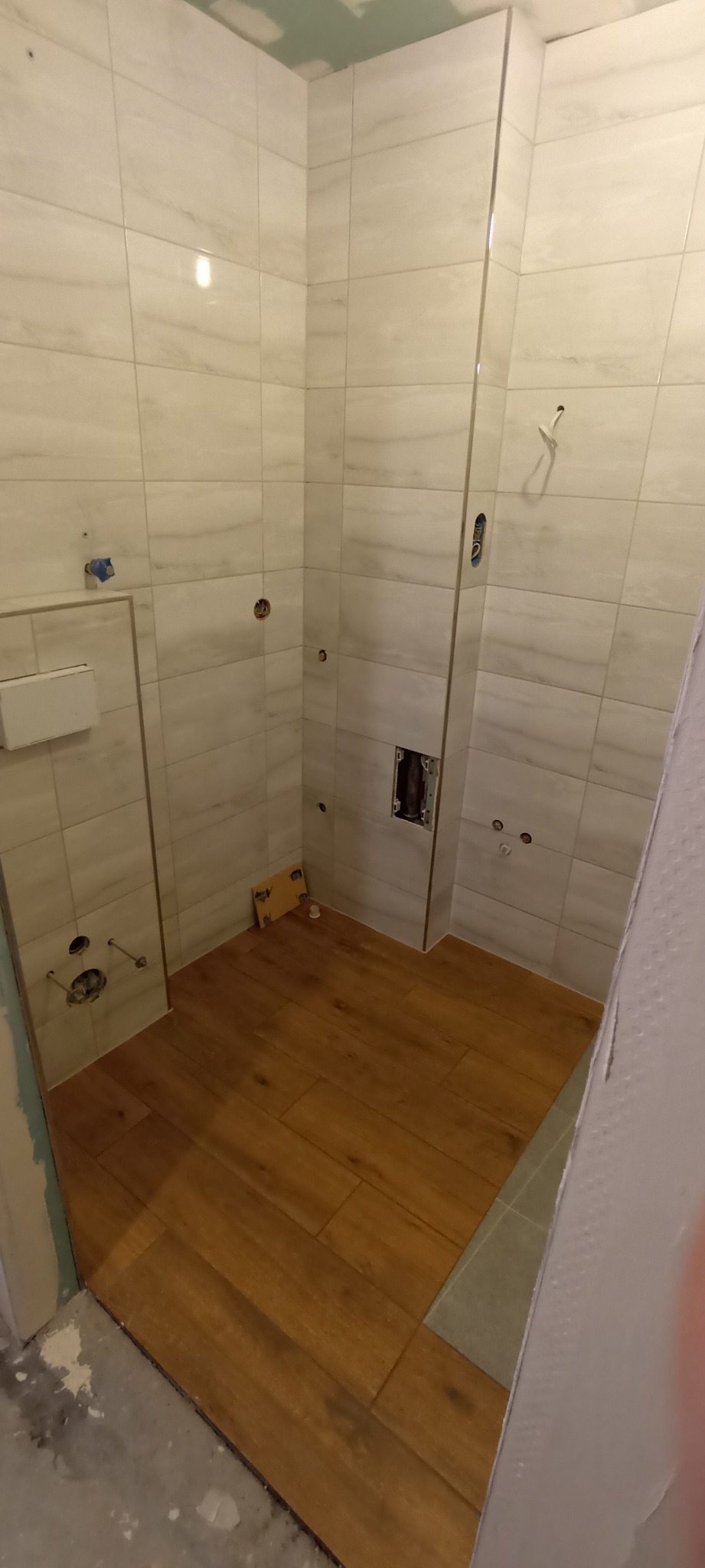 Niedokończona łazienka z ułożonymi panelami podłogowymi imitującymi drewno, białymi płytkami ściennymi z delikatnym marmurkowym wzorem, widocznymi otworami na armaturę i puszką elektryczną bez...