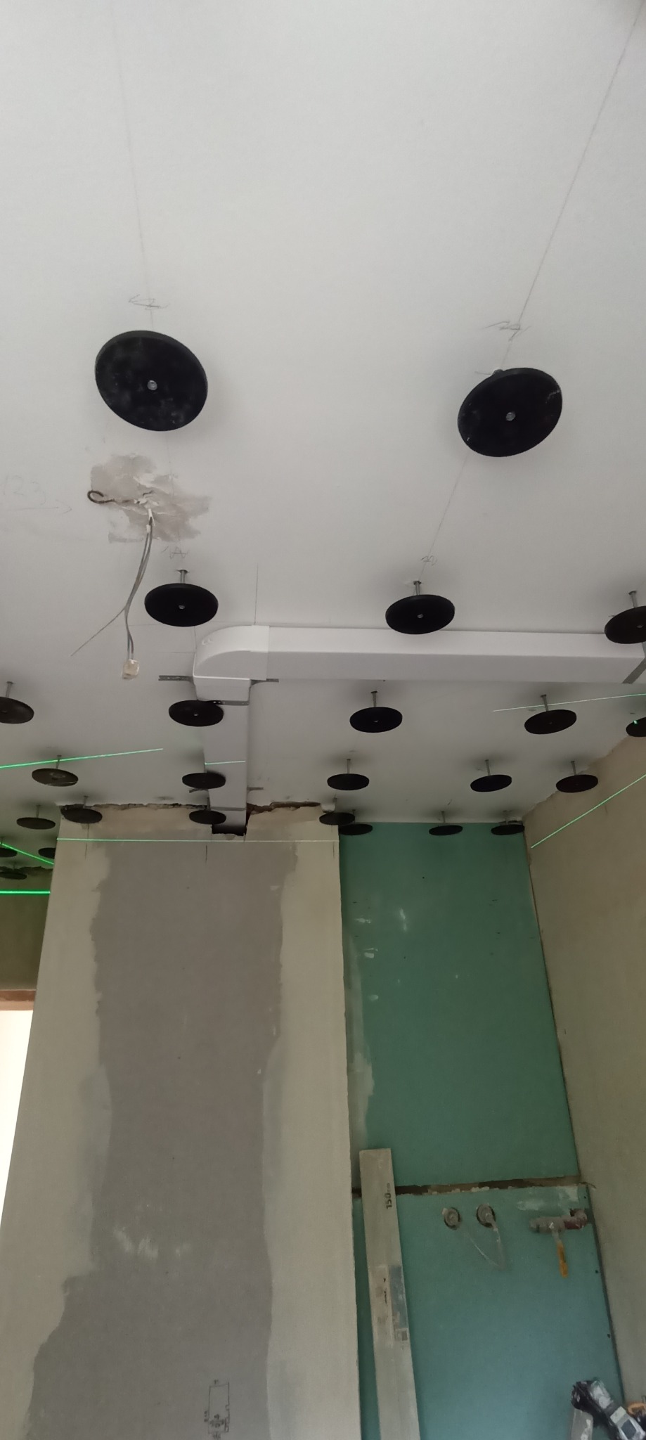 Montaż podwieszanego sufitu z płyt karton-gips, widoczne czarne uchwyty mocujące, laserowe wyznaczanie poziomu, częściowo wykończone ściany w kolorze szarym i zielonym w trakcie prac.