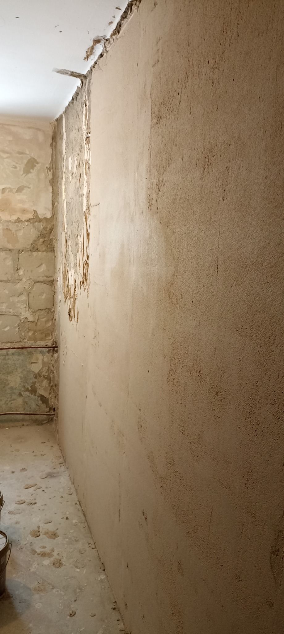 Ściana w trakcie prac przygotowawczych do wykończenia, widoczne fragmenty starego tynku, nałożona warstwa szpachli, odsłonięte cegły i instalacja elektryczna.