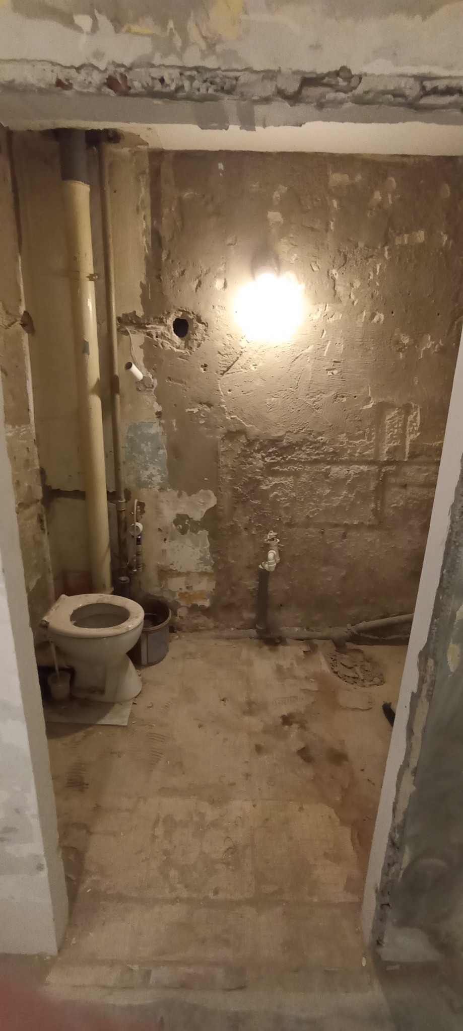 Łazienka w trakcie demontażu, widoczne stare płytki częściowo usunięte ze ścian, rury, stara toaleta i ślady po instalacjach.