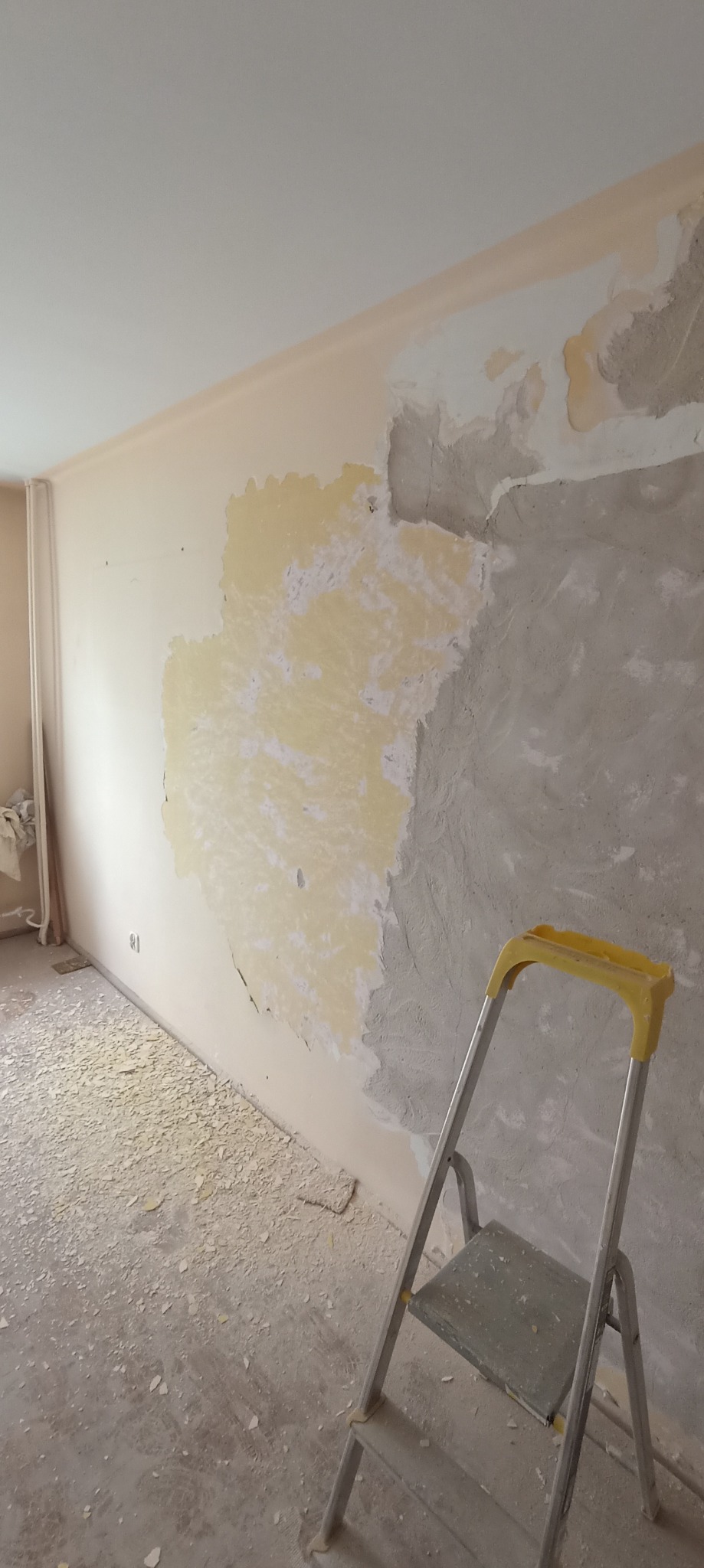 Ściana w trakcie przygotowania do malowania w Sanoczku, widoczne fragmenty starej farby, gładzi i szpachli, drabina malarska i resztki materiałów na podłodze.