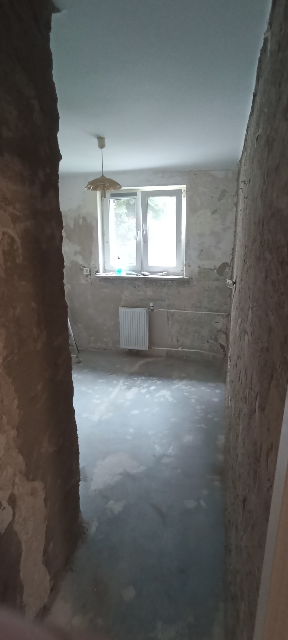 Pokój w trakcie przygotowania do prac wykończeniowych: surowe ściany z widocznymi warstwami, wylewka na podłodze, okno z białymi ramami i grzejnikiem pod spodem, wisząca lampa w stylu retro.