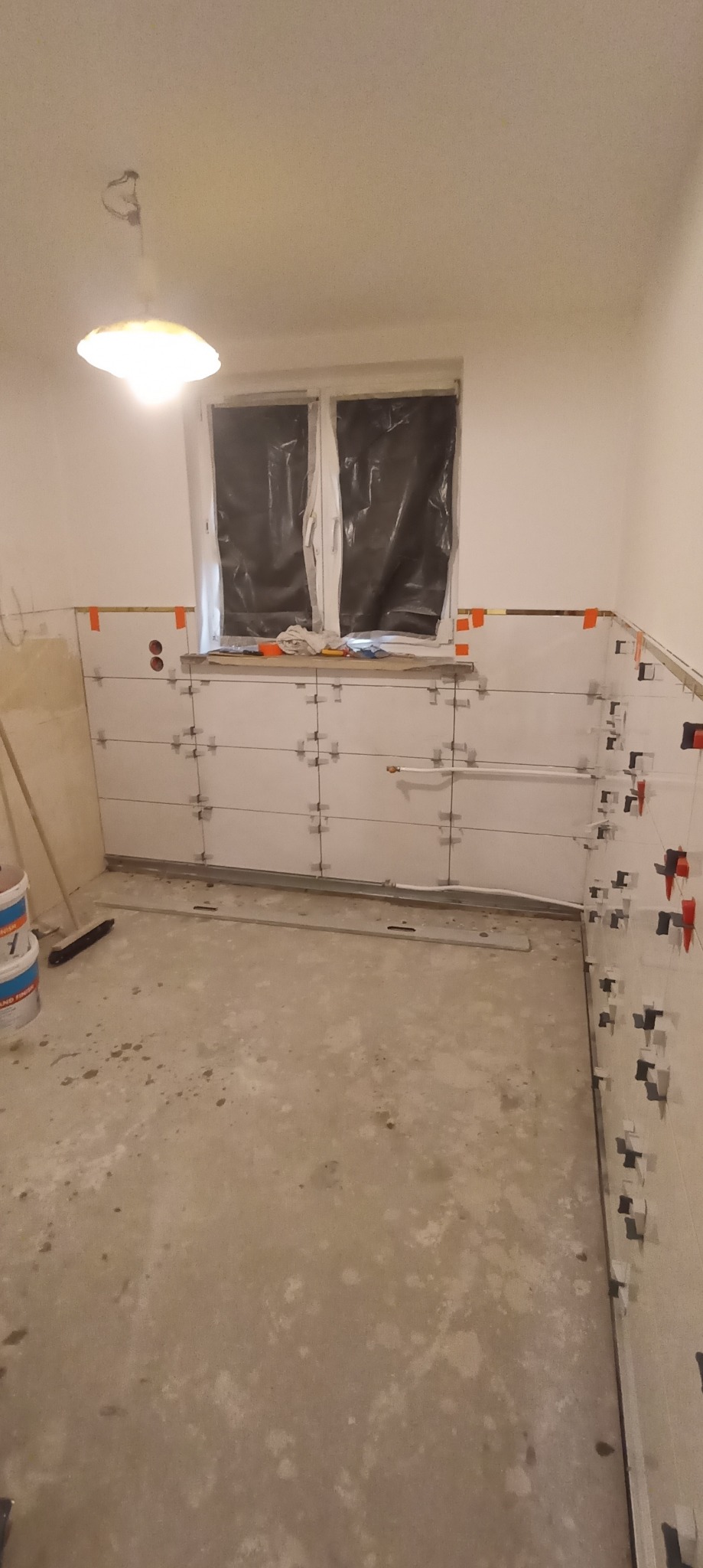 Świeżo ułożone, białe płytki ścienne w trakcie fugowania, z plastikowymi krzyżykami dystansowymi, widoczne narzędzia i poziomica, okno zasłonięte folią, surowy beton na podłodze.