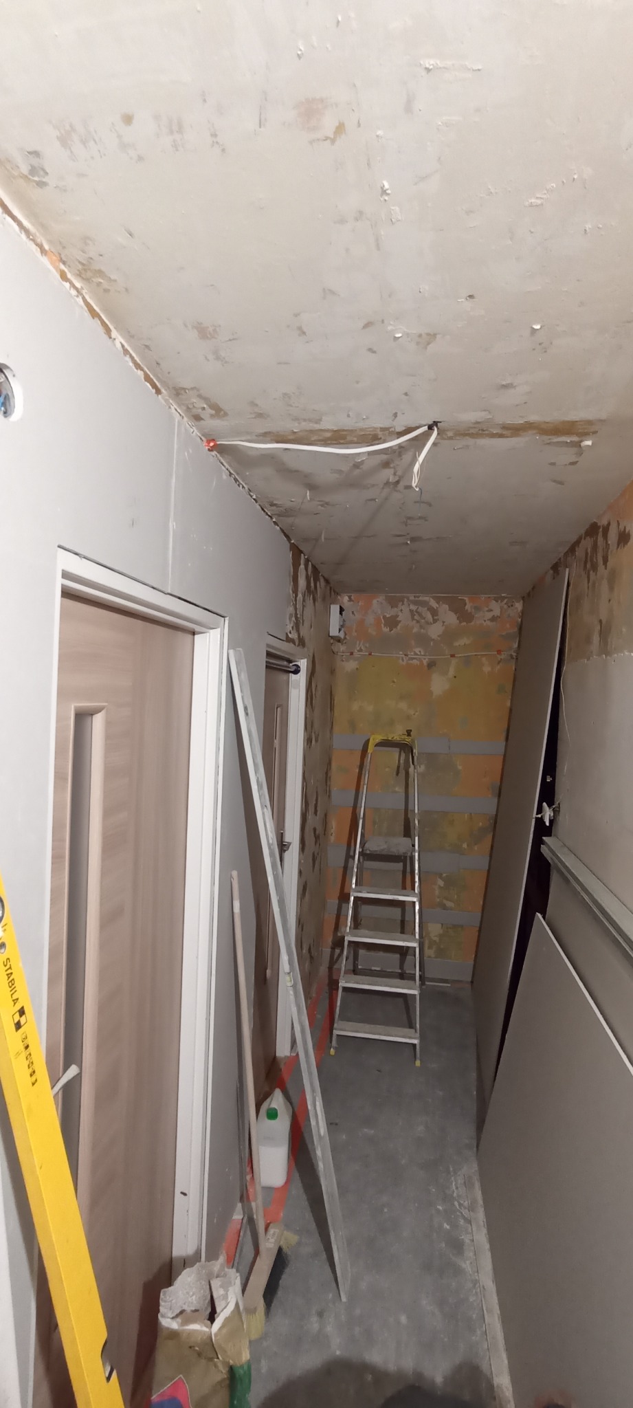 Remont korytarza w trakcie: ściany z płyt gips-karton, surowy sufit, drabina, narzędzia. Widok na korytarz w trakcie prac wykończeniowych.