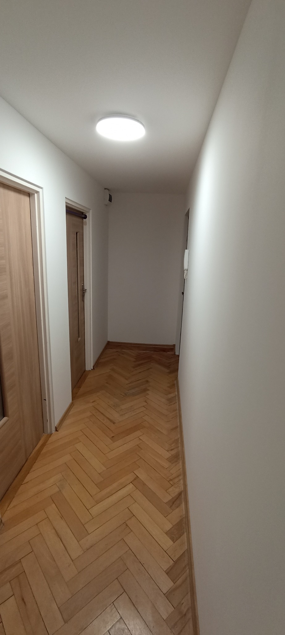 Korytarz po remoncie: parkiet ułożony w jodełkę, świeżo pomalowane białe ściany, widoczne drzwi i okrągła lampa sufitowa. Jasne, nowoczesne wykończenie.