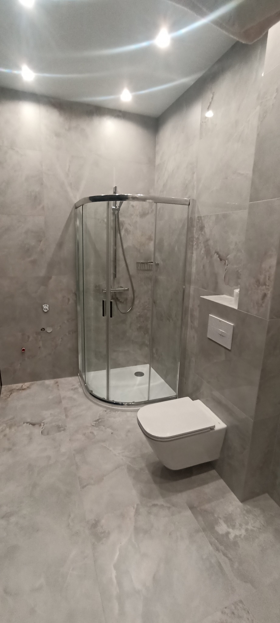 Nowoczesna łazienka w szarościach: kabina prysznicowa z chromowanymi detalami, podwieszana toaleta, płytki imitujące marmur na ścianach i podłodze, minimalistyczny design.