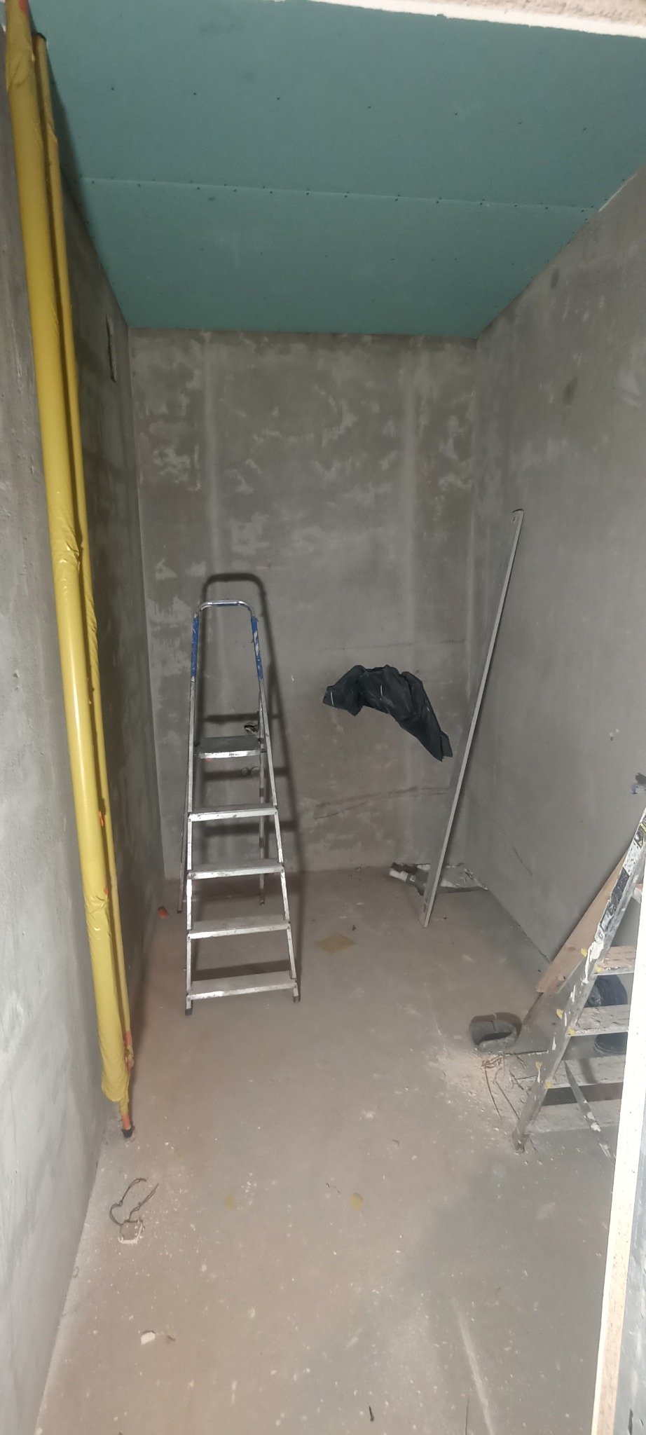 Wnętrze w trakcie prac: surowe ściany, zamontowana płyta karton gips na suficie. Dwie drabiny, rury w folii ochronnej. Widok na remontowany pokój.
