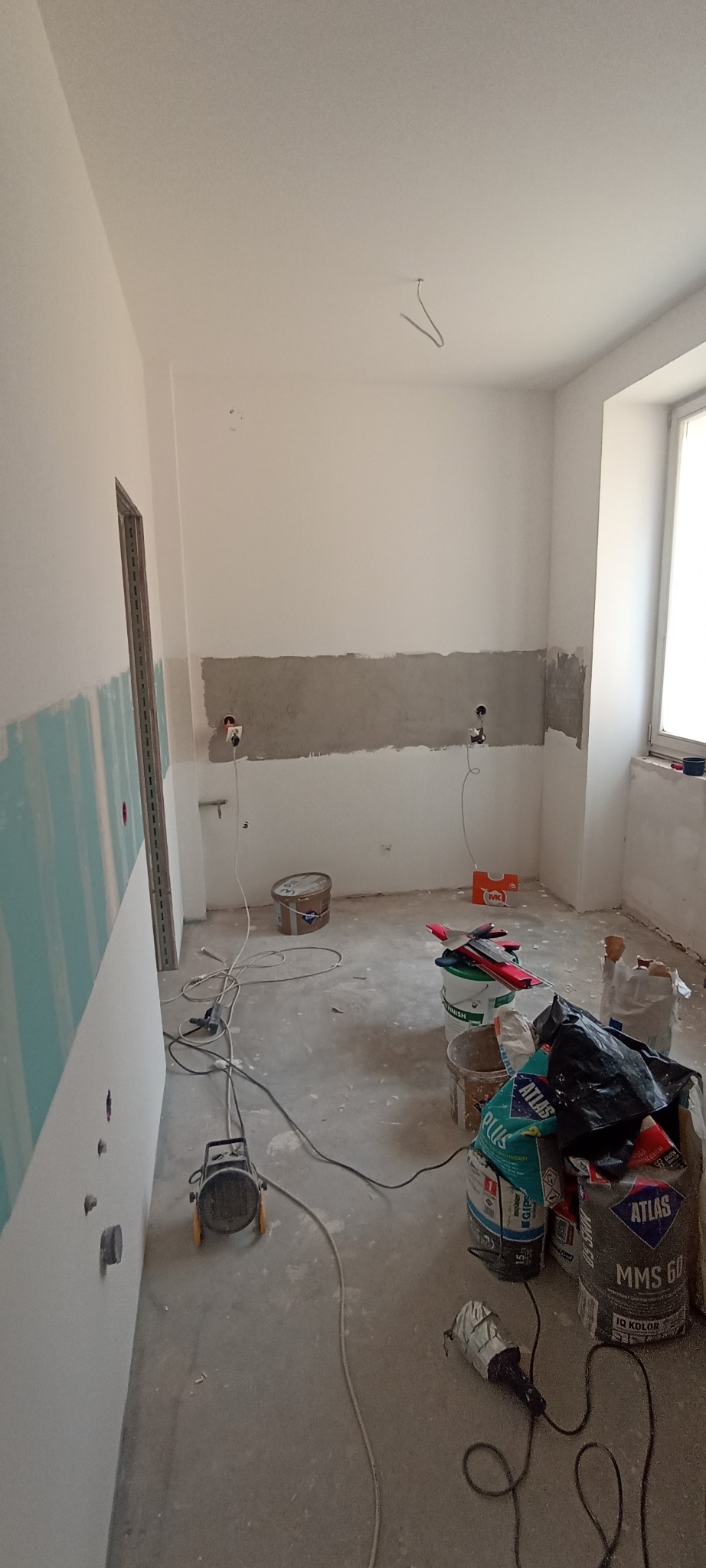 Wnętrze w trakcie prac: częściowo wyszpachlowana ściana, płyty gipsowe, worki z zaprawą, narzędzia. Widoczne przygotowania do dalszych prac wykończeniowych.