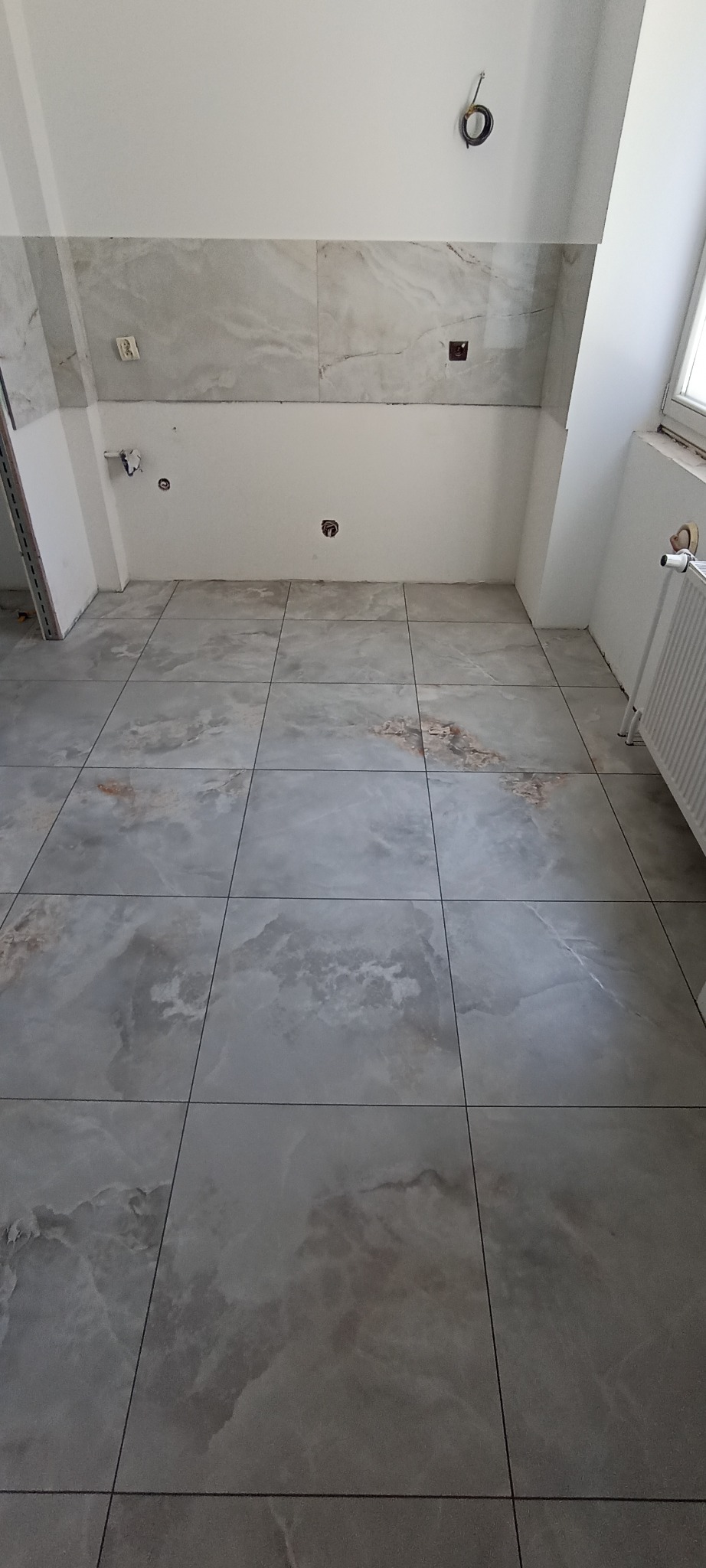 Wykończona łazienka z szarymi płytkami imitującymi marmur na podłodze i ścianach. Widoczne instalacje, grzejnik i fragment okna. Minimalistyczny styl.