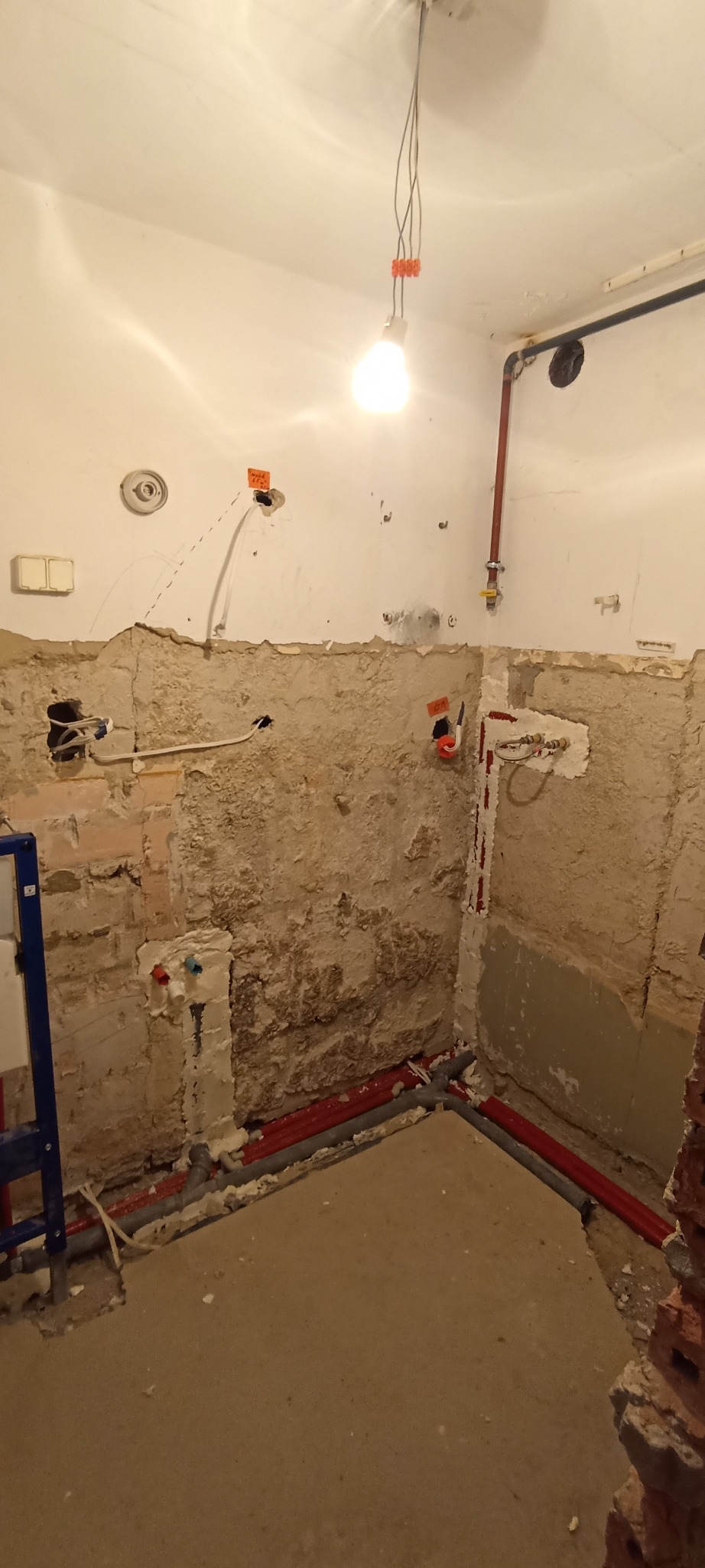 Wnętrze łazienki w trakcie remontu: odsłonięte instalacje wodne i elektryczne, surowe ściany z widoczną cegłą i resztkami tynku, przygotowane do dalszych prac wykończeniowych.