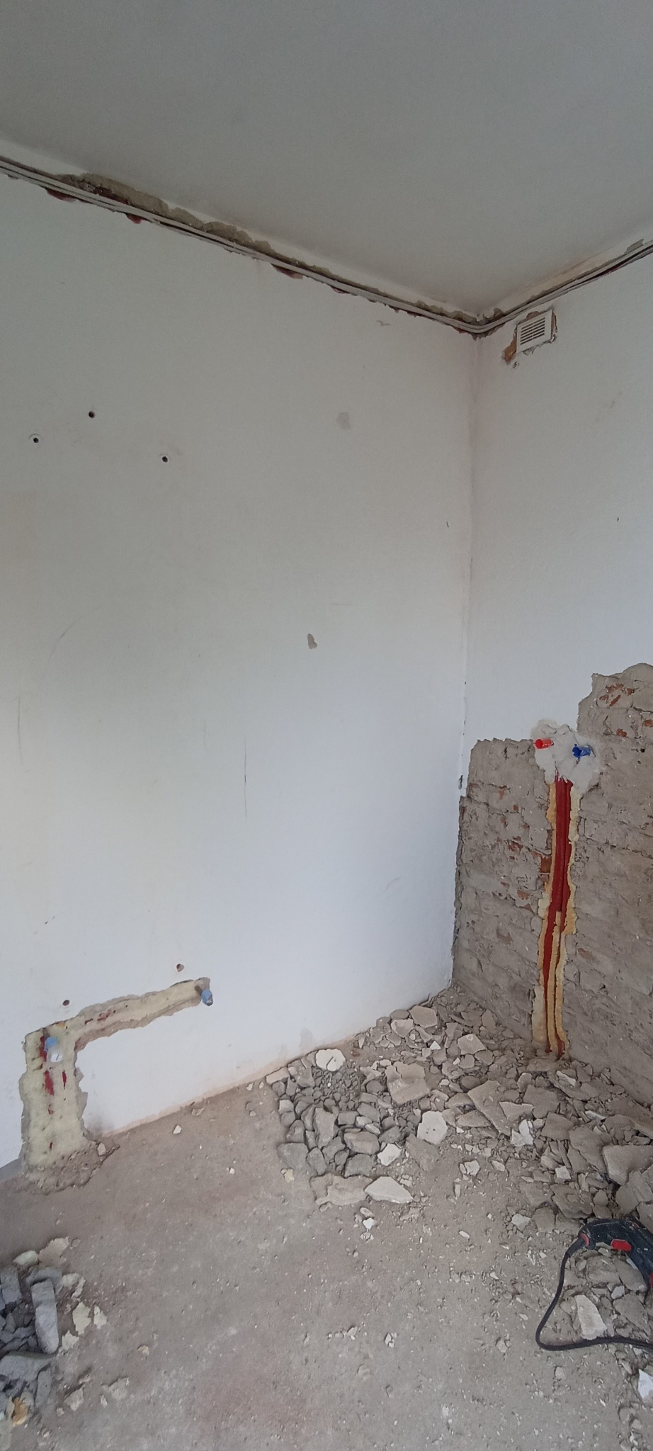 Fragment ściany z instalacją wodną w trakcie remontu. Widoczne rury w izolacji, gruz na podłodze i fragment narzędzia.