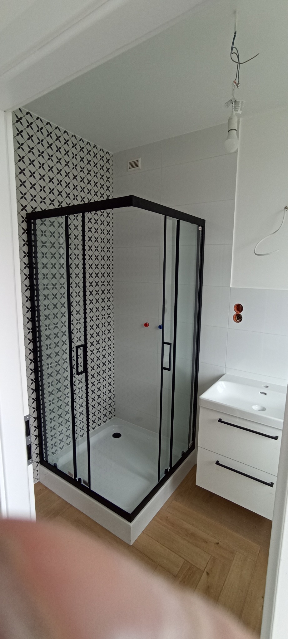 Nowoczesna kabina prysznicowa z czarnymi ramami i geometrycznym wzorem płytek w wykończonej łazience z białą szafką pod umywalką i drewnopodobną podłogą.