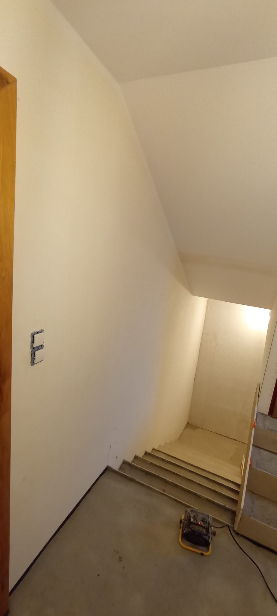 Klatka schodowa w trakcie remontu. Widoczne ściany po gładziowaniu, przygotowane schody i lampa budowlana. Na ścianie puszka elektryczna bez osprzętu.