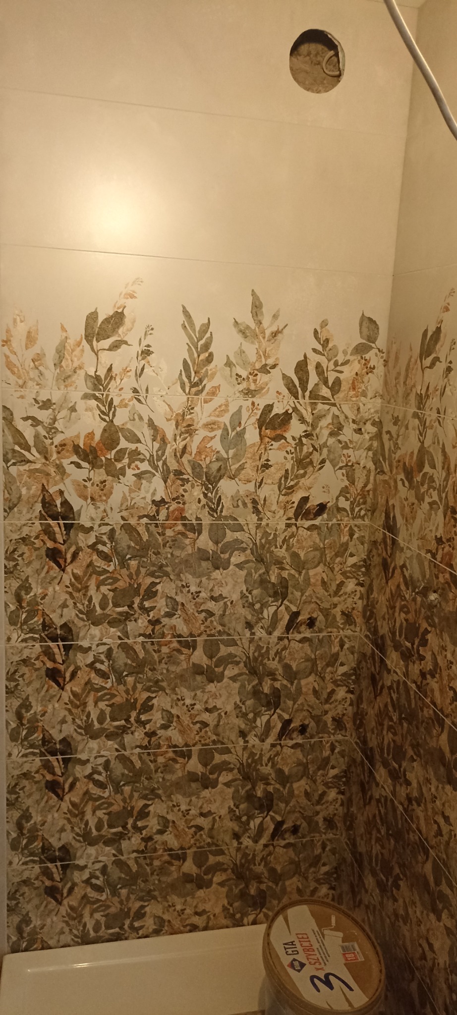 Wykończona ściana w łazience z dekoracyjnymi płytkami imitującymi liście. Widoczna puszka elektryczna bez osprzętu i wiadro z klejem do płytek w rogu pomieszczenia.