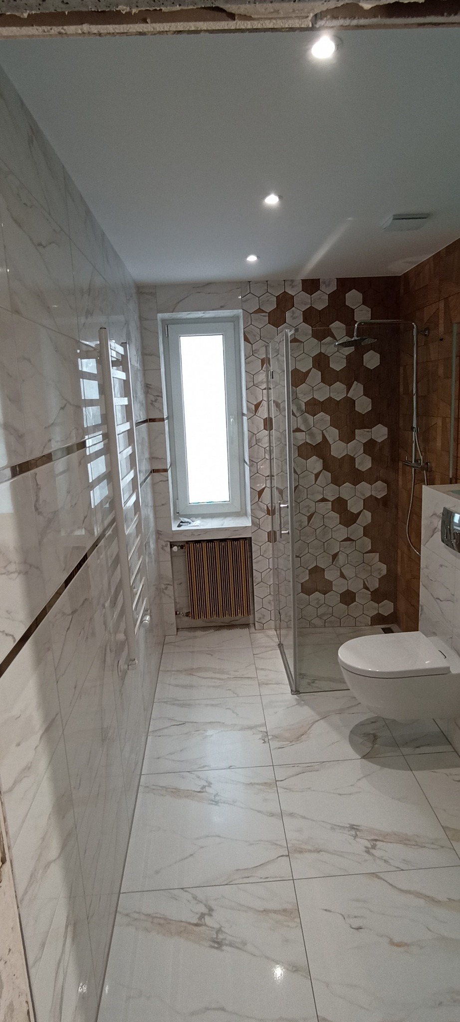 Wykończona łazienka z kabiną prysznicową, toaletą i grzejnikiem drabinkowym. Płytki imitujące marmur na ścianach i podłodze, dekoracyjne heksagony pod prysznicem.