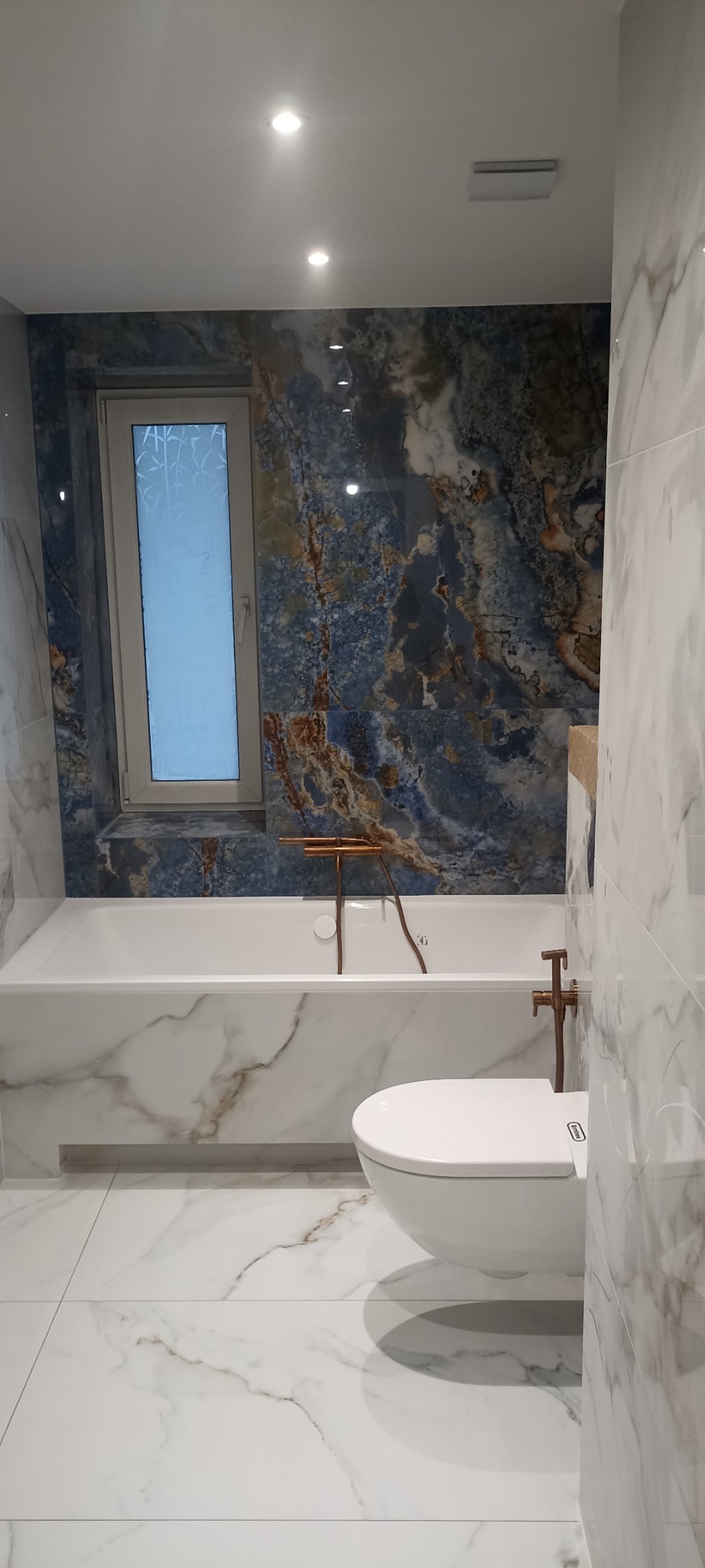 Elegancka łazienka z marmurowymi płytkami, białą wanną i toaletą. Ściana za wanną wykończona płytkami imitującymi niebieski marmur. Armatura w kolorze miedzi.