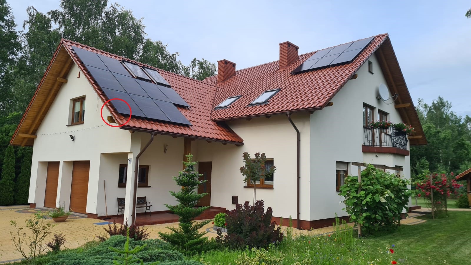 Dom jednorodzinny z panelami słonecznymi na dachu z czerwonej dachówki, widoczne dwa kominy i okna dachowe, ogród z krzewami i drzewami iglastymi.