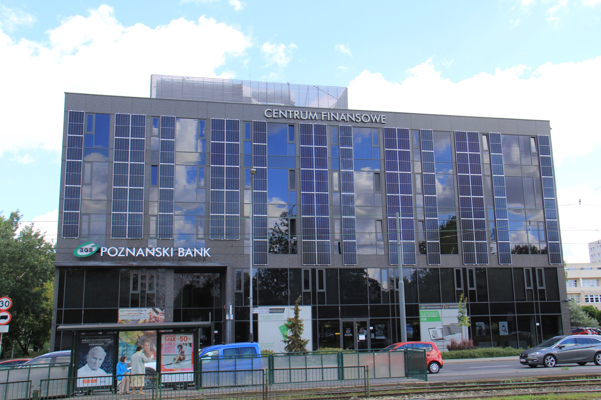 Budynek Centrum Finansowego Poznański Bank z elewacją częściowo pokrytą panelami słonecznymi, widok z poziomu ulicy, z przodu przystanek tramwajowy i przejeżdżające samochody.