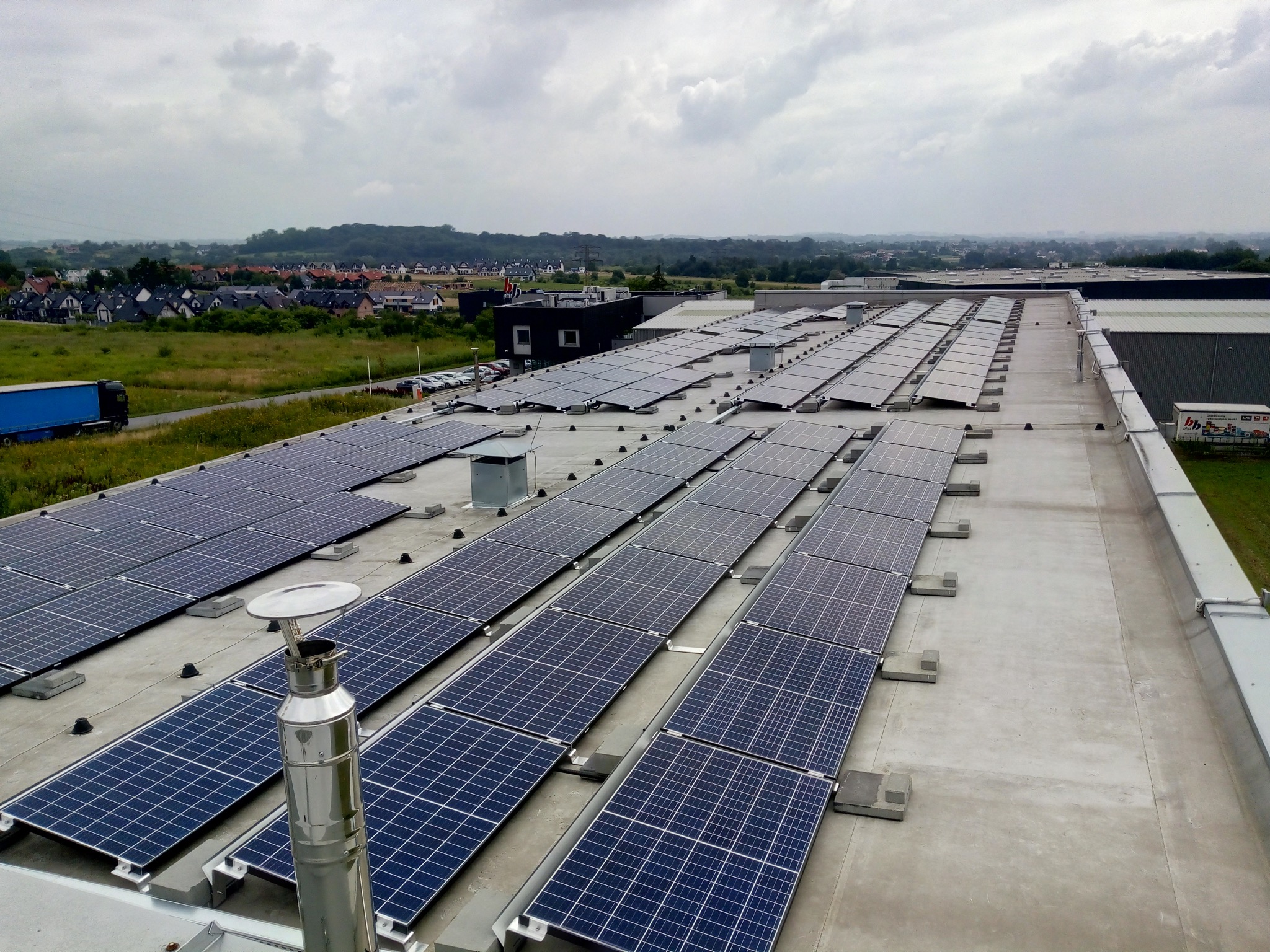 Rząd paneli fotowoltaicznych na płaskim dachu budynku przemysłowego, z kominem wentylacyjnym w pierwszym planie i widokiem na zabudowania mieszkalne w tle, pochmurne niebo.
