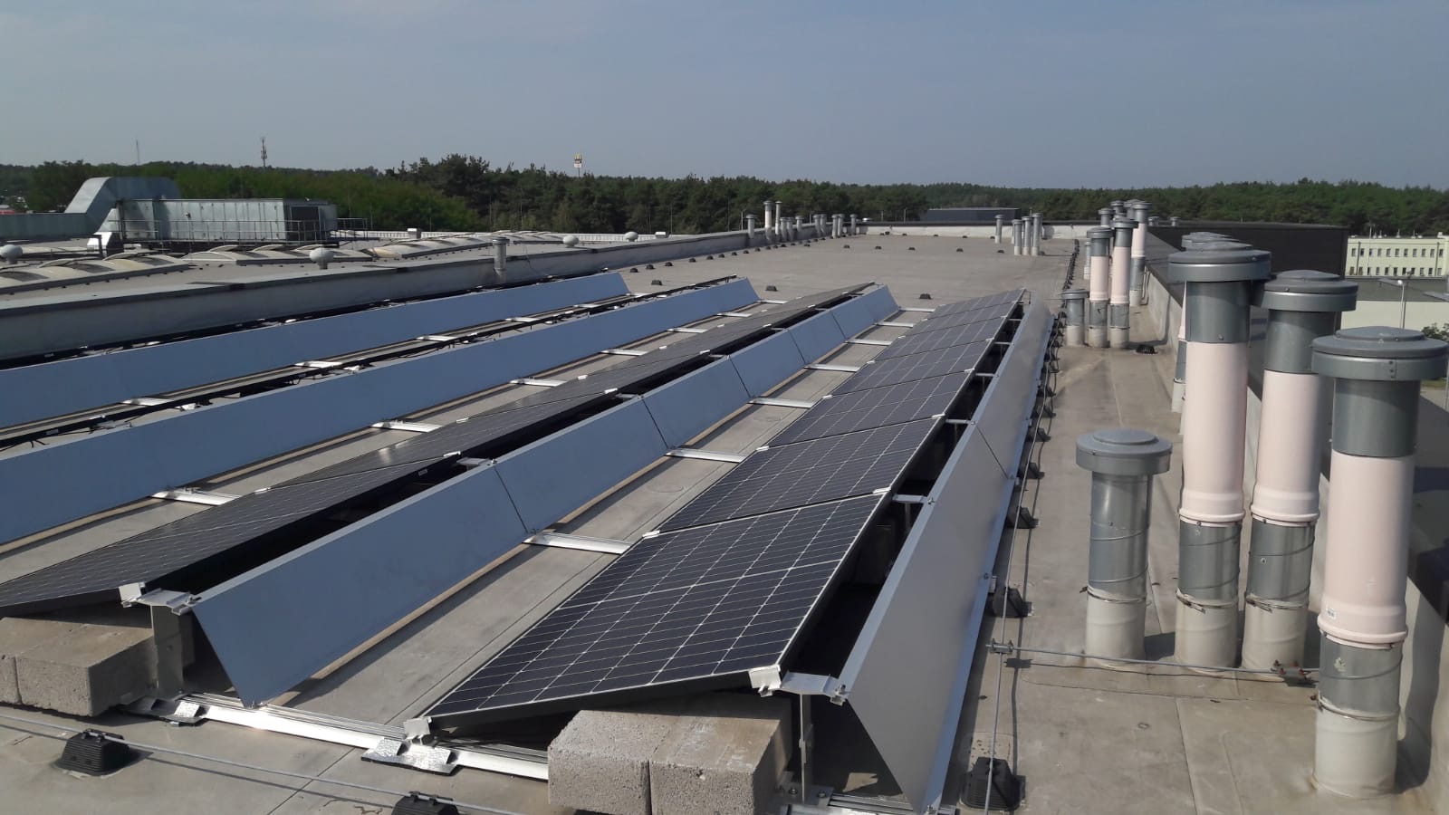 Panele słoneczne zamontowane na płaskim dachu budynku przemysłowego, widoczne szare osłony przeciwsłoneczne i kominy wentylacyjne w tle, drzewa w oddali.