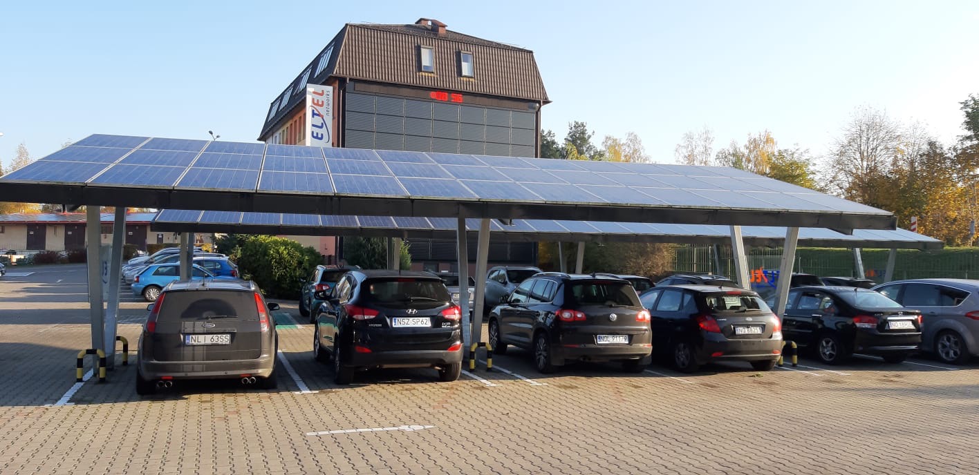 Zadaszony parking z panelami słonecznymi nad zaparkowanymi samochodami, budynek firmy ELTEL w tle.