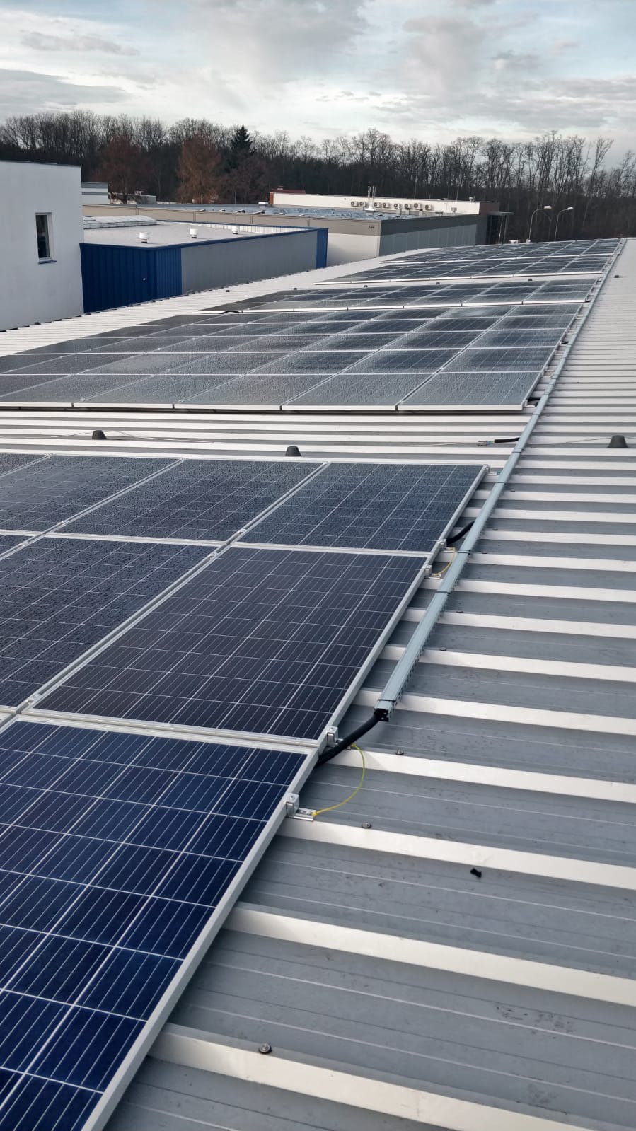 Rząd paneli słonecznych na metalowym dachu budynku przemysłowego, widoczne kable i elementy montażowe, pochmurne niebo w tle.