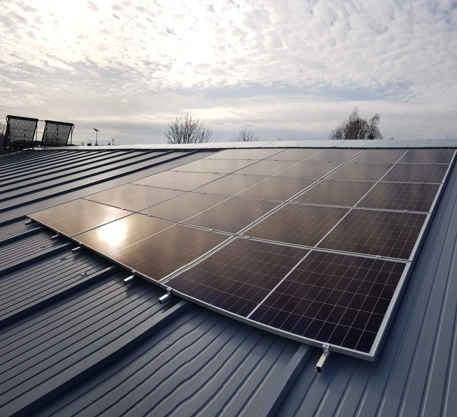 Panele słoneczne na metalowym dachu, z widocznymi kolektorami słonecznymi w tle pod pochmurnym niebem.