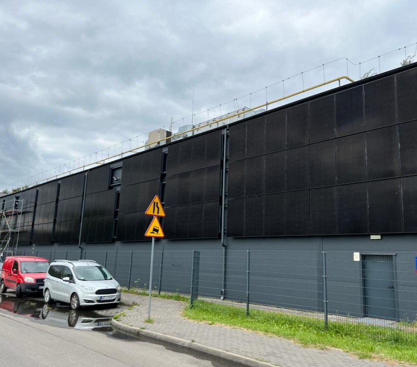 Duża instalacja paneli słonecznych na ścianie budynku przemysłowego, widoczne znaki drogowe i zaparkowane samochody, pochmurne niebo.