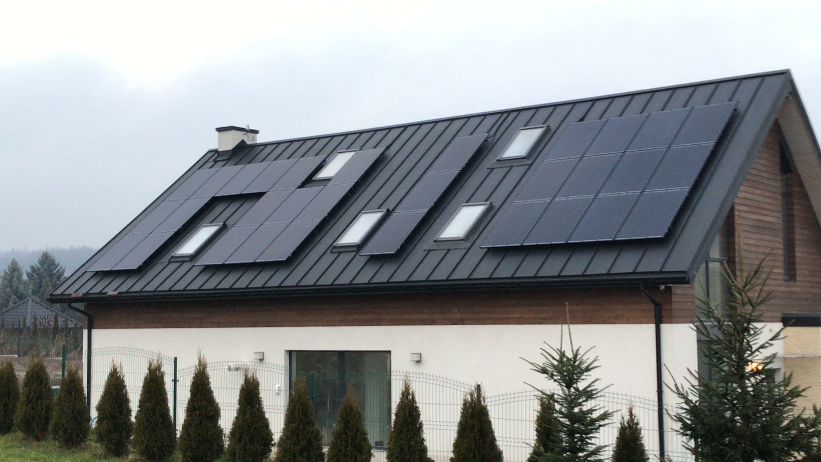 Czarny dach z panelami fotowoltaicznymi i oknami dachowymi na dwukondygnacyjnym domu z jasną elewacją i drewnianą ścianą szczytową, widoczne iglaki i metalowe ogrodzenie.