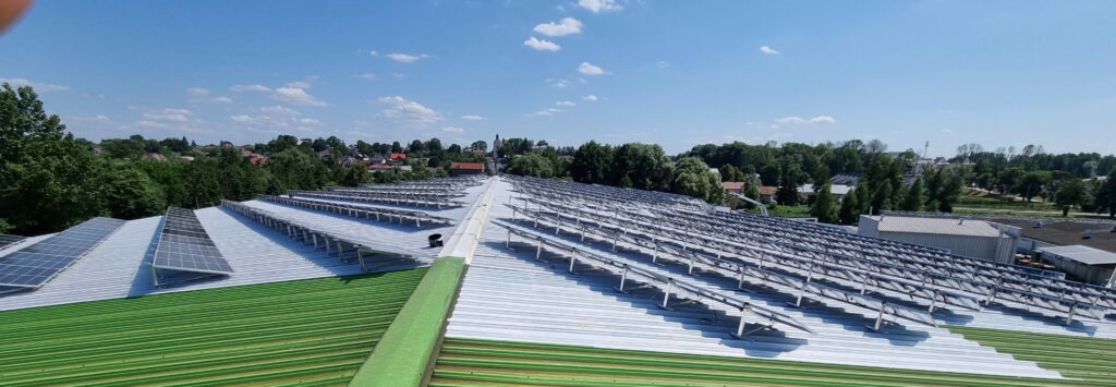 Rozległa instalacja paneli słonecznych na dachu przemysłowym z zielonymi elementami, widok z góry na tle zabudowań i zieleni miejskiej pod błękitnym niebem.