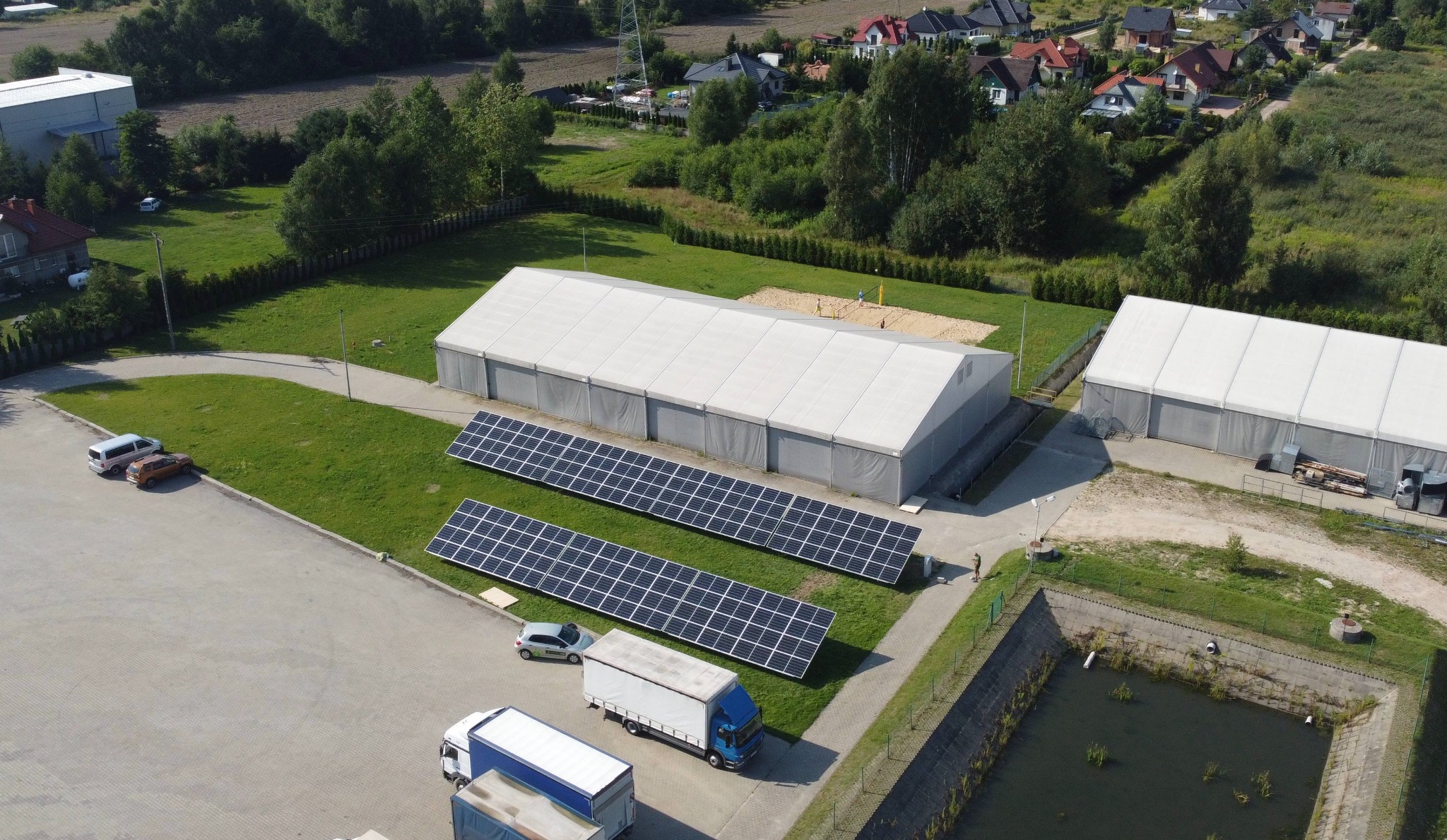 Widok z góry na instalację paneli słonecznych obok dużych namiotów magazynowych, na tle zieleni i zabudowań mieszkalnych.