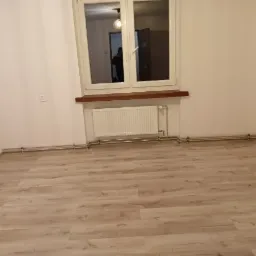 Pusty pokój po remoncie: jasne panele podłogowe, białe ściany, okno z drewnianym parapetem i grzejnik. Widoczna instalacja grzewcza przy podłodze.