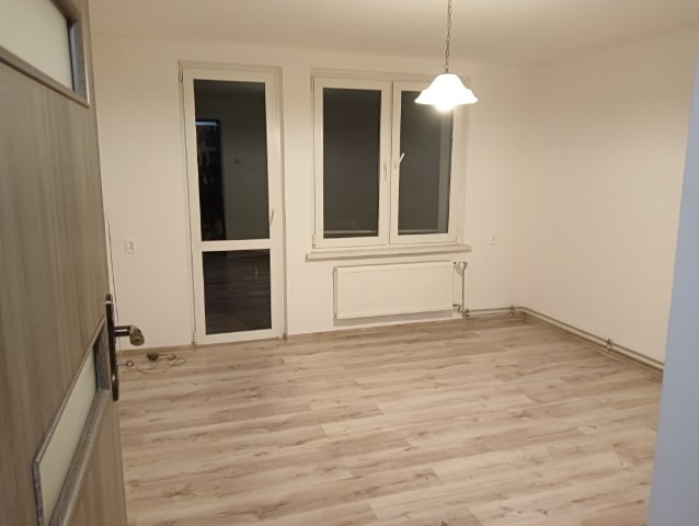Pusty pokój po remoncie z panelami podłogowymi, białymi ścianami, oknem i drzwiami z widocznym grzejnikiem i lampą wiszącą, uchwyt drzwiowy w lewym dolnym rogu.