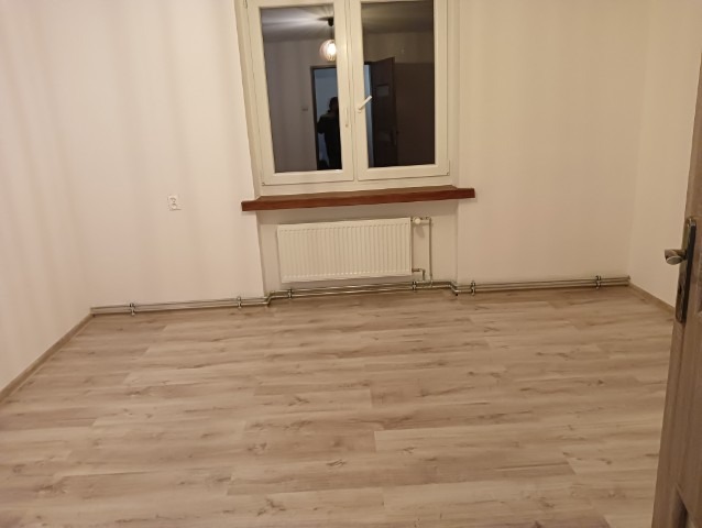 Pusty pokój po remoncie: jasne panele podłogowe, białe ściany, okno z drewnianym parapetem i grzejnik. Widoczna instalacja grzewcza przy podłodze.