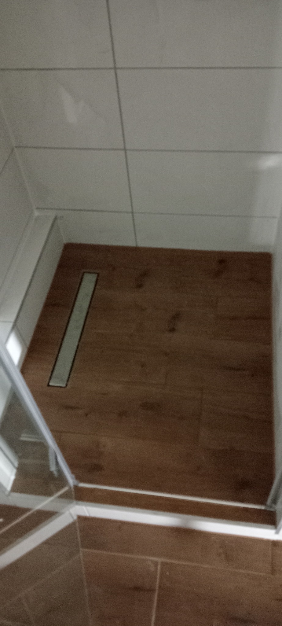 Nowoczesny odpływ liniowy w kabinie prysznicowej z drewnopodobną podłogą i jasnymi, kwadratowymi płytkami na ścianach.