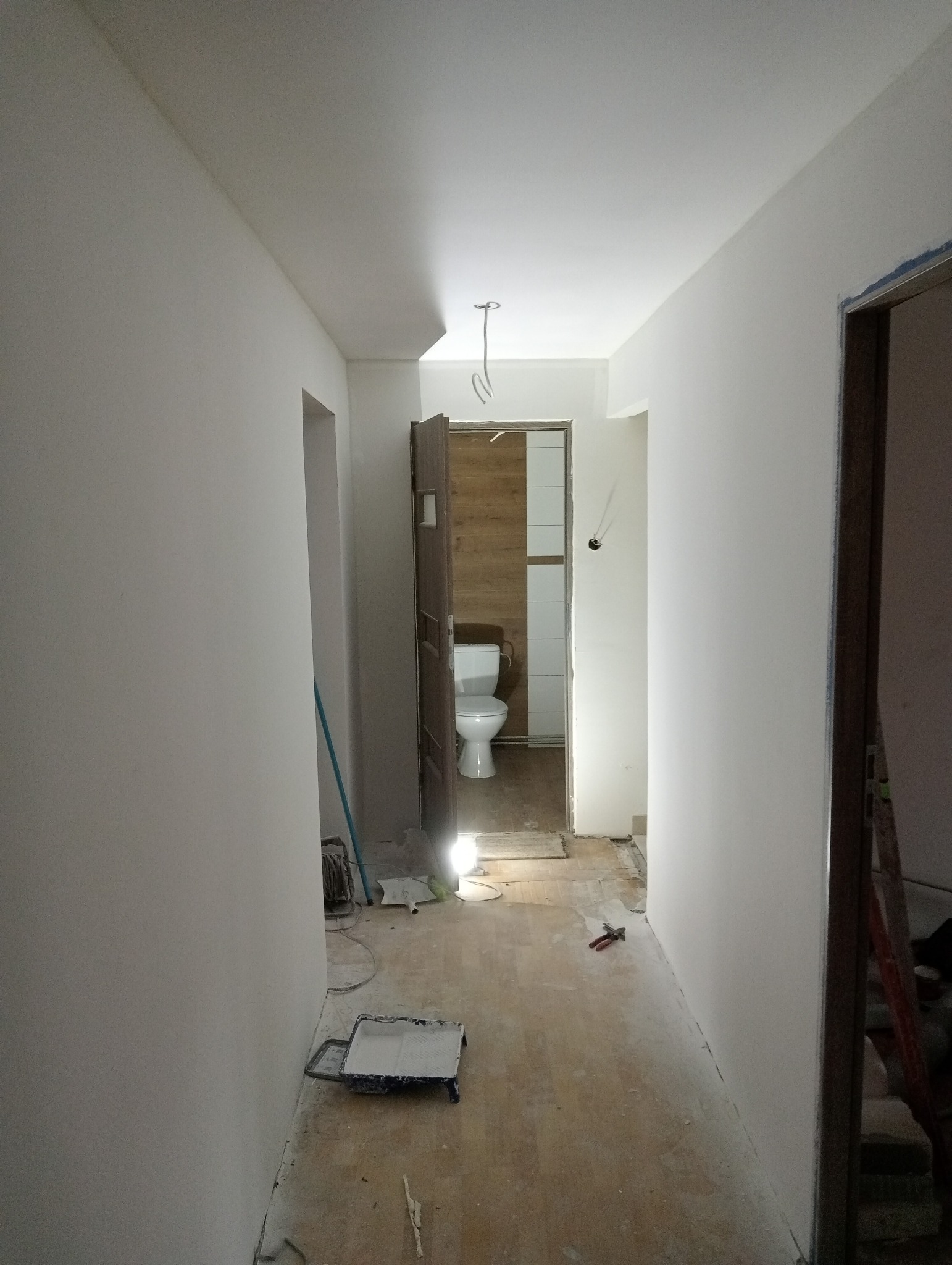 Korytarz w trakcie malowania na biało, widoczny otwarty dostęp do łazienki z drewnianymi panelami i białą toaletą, kuweta malarska na podłodze.