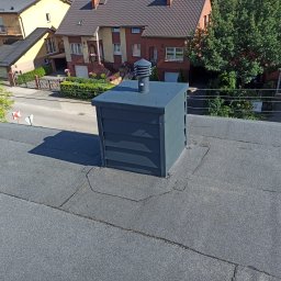 Janusz Bach - Widok z góry na płaski dach pokryty papą z zainstalowanym, nowym kominem wentylacyjnym w kolorze grafitowym; w tle budynki mieszkalne i ulica.