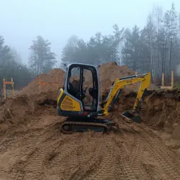 Żółto-szara mini koparka Wacker Neuson ET18 na dnie wykopu pod fundamenty, widoczne ślady gąsienic, drewniane szalunki i mglisty las w tle.