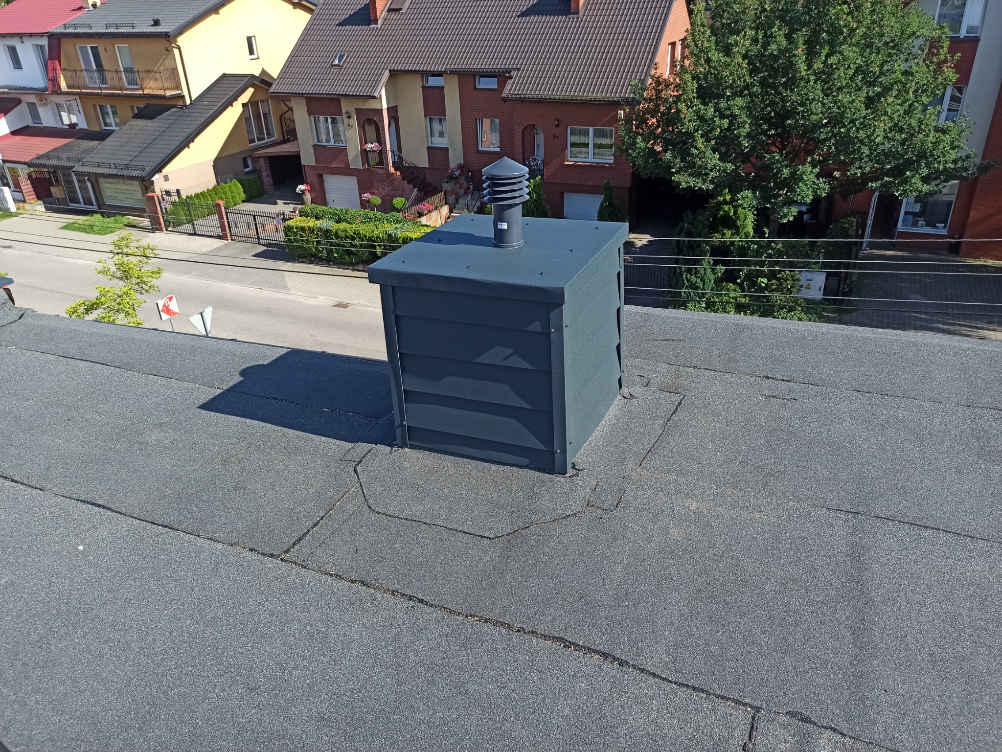 Widok z góry na płaski dach pokryty papą z zainstalowanym, nowym kominem wentylacyjnym w kolorze grafitowym; w tle budynki mieszkalne i ulica.